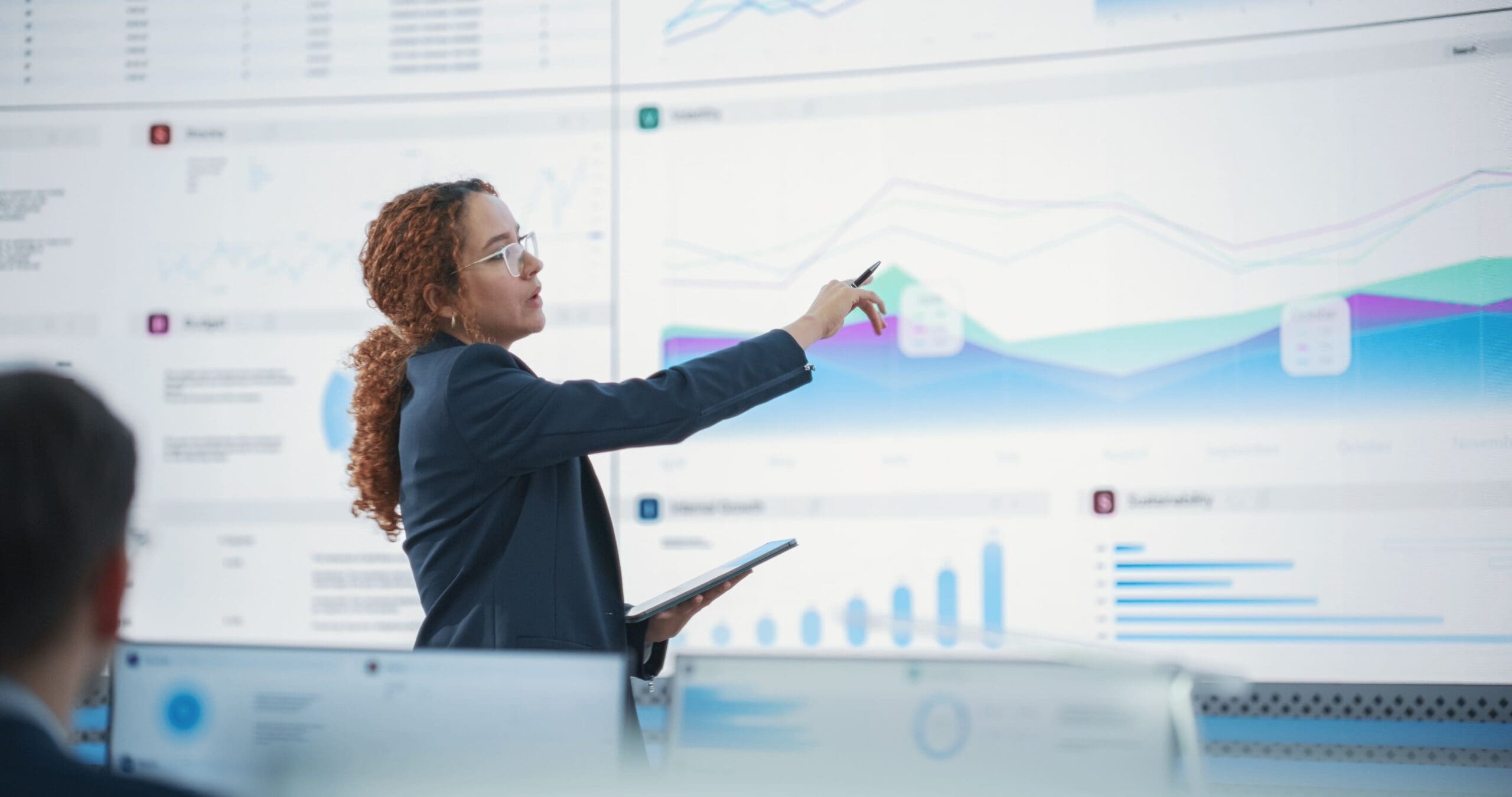 Female Senior Data Scientist Reviewing Reports On Big Digital Screen In Monitoring Room. Diverse Consulting Company Employees Working Behind Desktop Computers.